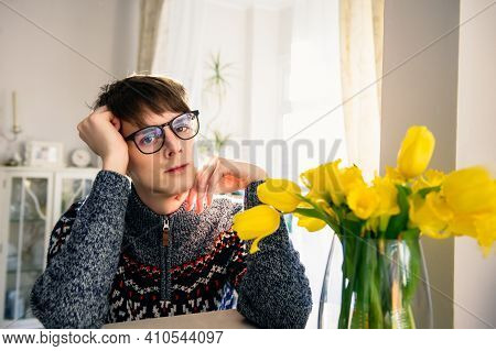 Portrait Of Young Sensual Teenager Man Seating At The Table With Yellow Flowers Bouquet On Light Int