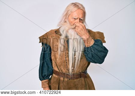 Old senior man with grey hair and long beard wearing viking traditional costume smelling something stinky and disgusting, intolerable smell, holding breath with fingers on nose. bad smell 