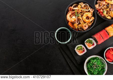Assorted sushi rolls and wok noodles with shrimps and vegetables in a black bowl, seaweed salad on a black slate background, top view, copy space. Traditional Asian food set.