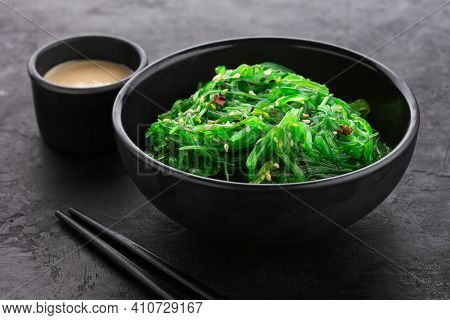 Hiyashi Wakame Chuka seaweed salad with sesame seeds in a black bowl and peanut sauce next to it on a black textured background.