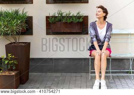 Young Adult Beautiful Caucasian Woman Sitting On Metal Vintage Bench Chair Against Wall With Many Mo
