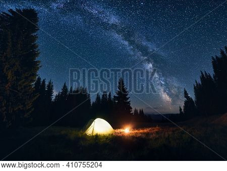 Panoramic View Of Night Camping In Valley With Large Pine Trees. Burning Campfire And Illuminated To
