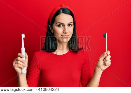 Young hispanic woman choosing electric toothbrush or normal teethbrush relaxed with serious expression on face. simple and natural looking at the camera. 