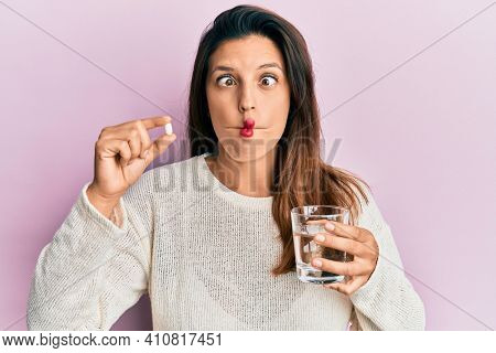 Beautiful hispanic woman holding pill and glass of water making fish face with mouth and squinting eyes, crazy and comical. 