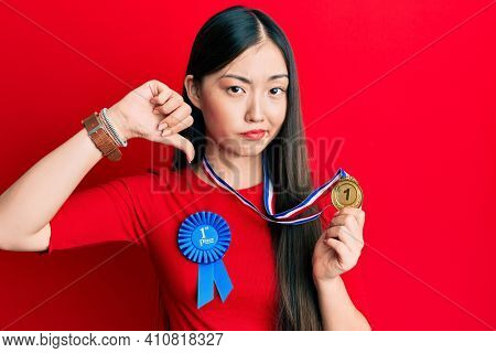 Young chinese woman wearing first place badge holding medal with angry face, negative sign showing dislike with thumbs down, rejection concept 