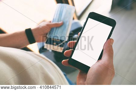 The man holding the mobile phone with empty space as a mockup for digital vaccination certificate in one hand and passport, mask and ticket in another one