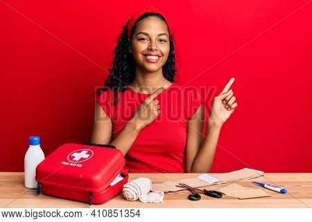 Young african american girl sitting on the table with first aid kit making cure smiling and looking at the camera pointing with two hands and fingers to the side. 