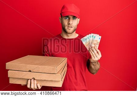 Young caucasian man holding take away food and 10 euros banknotes in shock face, looking skeptical and sarcastic, surprised with open mouth 