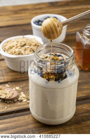 Yogurt, Granola, Bluberry And Honey Over Wooden Table. Wood Utensil Pouring Honey.