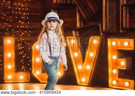 Portrait Of A Little Girl Wearing A Hat On A Background Of Wooden Letters Decorated With Lamps