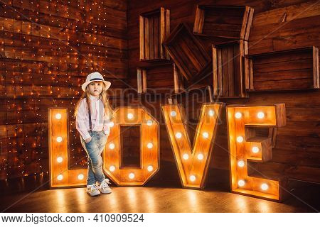 A Little Girl Wearing A Hat Stands Against A Background Of Wooden Letters Decorated With Lamps