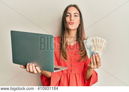 Young beautiful woman holding laptop and denmark krone looking at the camera blowing a kiss being lovely and sexy. love expression. 