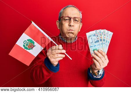 Handsome senior man with grey hair holding peru flag and peruvian sol banknotes puffing cheeks with funny face. mouth inflated with air, catching air. 