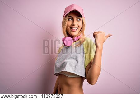 Young beautiful blonde sportswoman doing sport using headphones over pink background smiling with happy face looking and pointing to the side with thumb up.