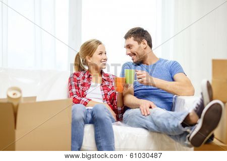 moving, home and couple concept - smiling couple with coffee or tea cups relaxing on sofa in new home