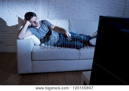 young attractive man at home lying on couch at living room watching tv holding remote control and changing channel or volume looking bored