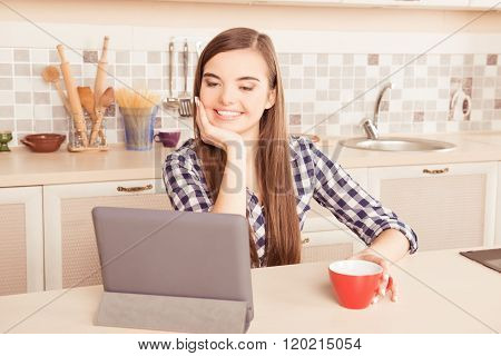Young Girl Sittiing In The Kitchen With Tea And Reading News On Tablet