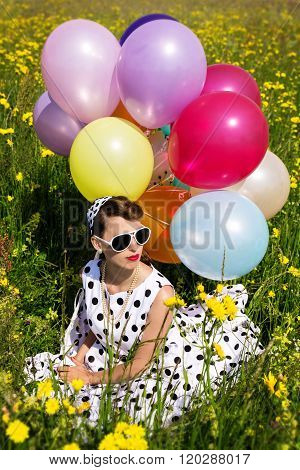 Beautiful Woman With A Rockabilly Style Dress Sitting On A Flowery Meadow With Colorful Balloons