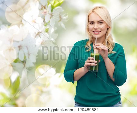 healthy eating, vegetarian food, diet, detox and people concept - smiling young woman drinking green vegetable juice or smoothie from glass over natural cherry blossom background