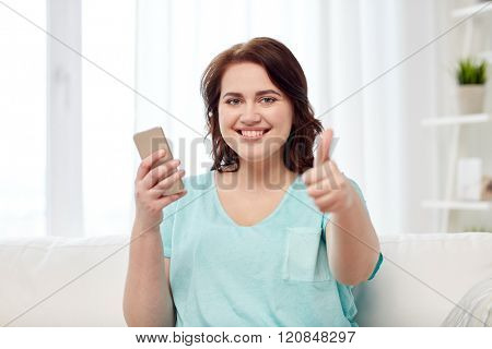 people, technology, communication and leisure concept - happy young plus size woman sitting on sofa with smartphone at home and showing thumbs up