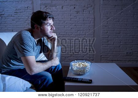 young happy television addict man sitting on home sofa watching TV and eating popcorn looking mesmerized smiling while enjoying movie sitcom or live sport at night