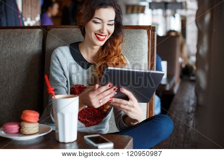 A bright ,beautiful woman with curly red hair,red lipstick,beautiful makeup,red nail Polish,sitting alone at a table in a cafe with a Cup of coffee latte and vanilla cookies,working with tablet computer in the Internet