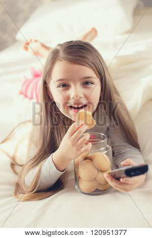 Cute little 6 years old girl,brunette with long flowing hair,dressed in a gray t-shirt and pink striped pants,spends his time at home,lying on white bed near the window with the TV control in one hand,eating oatmeal cookies from the glass jar