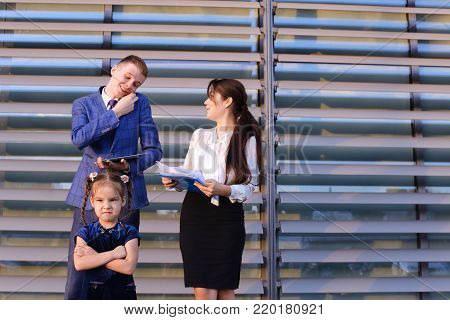 In  foreground stands and frowns, angry with arms crossed in front of little girl, child,  in background successful young handsome man's man, young father and modern woman, young mother hold gadgets in hand, mobile phones and talking to them, solve operat