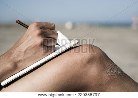 closeup of a young caucasian man writing with a pen in a notebook on his shaved legs, on the beach during the summer