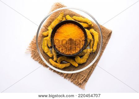Turmeric powder in ceramic bowl with raw dried turmeric over plain background