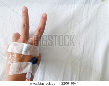 Patient hand with V sign and the tube of normal saline infusion on white cloth background (Victory for the disease)