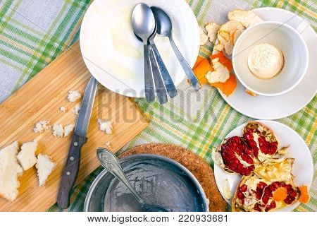 Messthetics concept. An overhead photo of dirty bowls with oatmeal porridge leftovers, half of pomegranate, tea spoons and knife, empty saucepan, wooden chopping board and a cup of espresso coffee