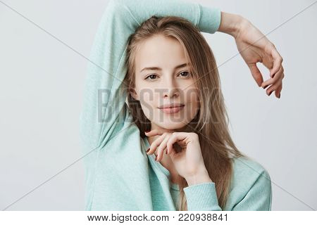 Headshot of happy pleased blonde girl of European appearance with dark eyes wearing blue long-sleeved top looking and smiling at camera, standing isolated against gray background. Positive emotions and feelings.