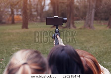 Rear View Of Three Girlfriends Sitting On The Grass In A Park Taking A Selfie Using A Gimbal