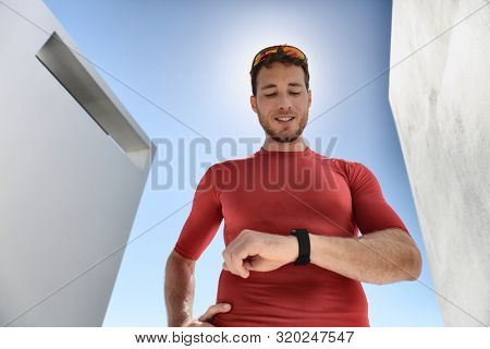 Athlete runner checking cardio on sports smartwatch jogging on outdoor run track. Running man wearing sunglasses and tech wearable device looking at watch during training workout.