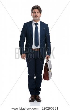 Confident businessman holding a suitcase and looking forward while wearing an elegant blue suti and tie, standing on white studio background