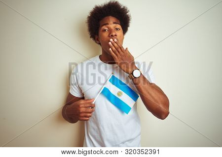 Afro american tourist man holding Argentinian Argentina flag over isolated white background cover mouth with hand shocked with shame for mistake, expression of fear, scared in silence, secret concept