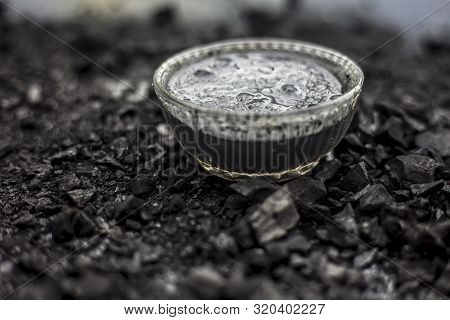 Close Up Of Activated Charcoal In A Glass Bowl On The Wooden Surface Along With Some Raw Powder Of C
