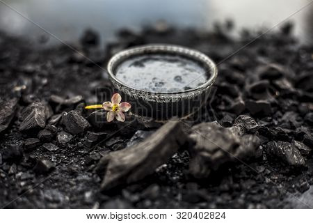 Close Up Of Activated Charcoal In A Glass Bowl On The Wooden Surface Along With Some Raw Powder Of C