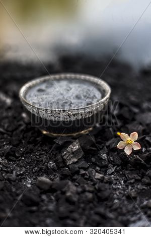 Close Up Of Activated Charcoal In A Glass Bowl On The Wooden Surface Along With Some Raw Powder Of C