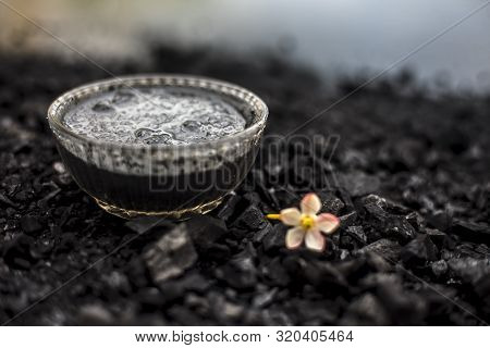 Close Up Of Activated Charcoal In A Glass Bowl On The Wooden Surface Along With Some Raw Powder Of C