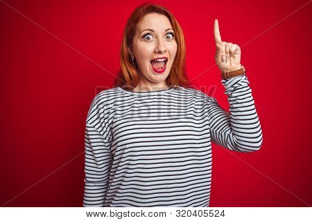 Young redhead woman wearing strapes navy shirt standing over red isolated background pointing finger up with successful idea. Exited and happy. Number one.