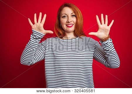 Young redhead woman wearing strapes navy shirt standing over red isolated background showing and pointing up with fingers number ten while smiling confident and happy.
