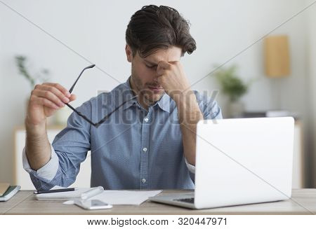 Overworked Guy Massaging Nosebridge Holding Eyeglasses Sitting At Laptop Indoor. Migraine And Eyes F