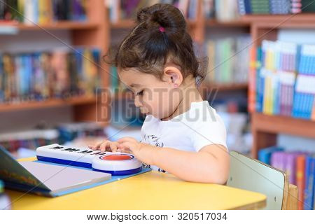 Little Child Indoors In Front Of Books. Cute Young Toddler Sitting On A Chair Near Table And Play Wi