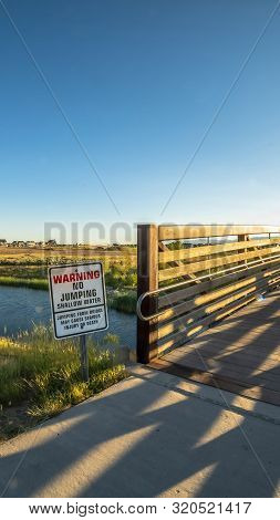 Vertical Frame Warning Sign And Bridge Over A Lake Sourrounded By Houses And Grassy Terrrain