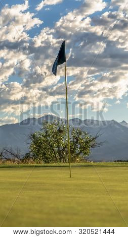 Vertical Frame Focus On The Flagstick Of A Golf Course Against Trees Mountain And Cloudy Sky