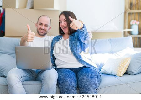 Young couple sitting on the sofa arround cardboard boxes moving to a new house using laptop happy with big smile doing ok sign, thumb up with fingers, excellent sign