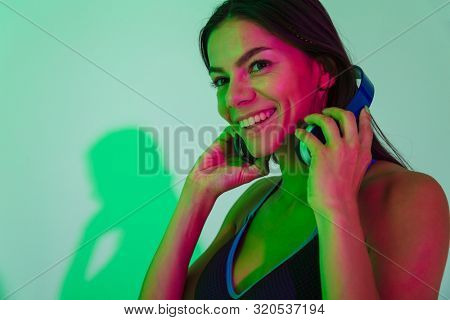 Image of happy smiling optimistic young strong fitness woman isolated with led flash lights listening music with headphones.