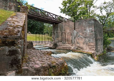 Wooden Bridge Over The River Vilia In Vilnius With A Waterfall In The Park Belmonto.